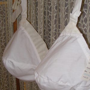 May include: A white bra with soft, unpadded cups and adjustable shoulder straps. The garment features a tag with the text M1080, WHITE, and RESTFORM. The bra is suspended on a plastic hanger, showcasing a simple, functional design.