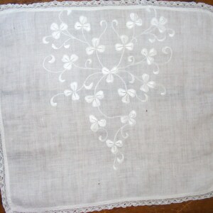 May include: White handkerchief with a delicate floral embroidery design. The embroidery features a stylized clover pattern. The edges are finished with a delicate lace trim. The handkerchief is square in shape.