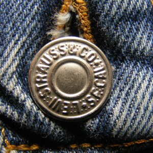 May include: Close-up of a silver button from a pair of blue denim jeans. The button is embossed with the words "STRAUSS & CO. / S.F. CAL." and "LEVI'S" around the edge. The denim fabric is a classic blue with visible stitching.