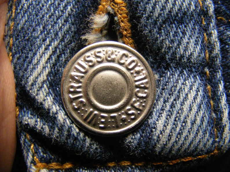 May include: Close-up of a silver button from a pair of blue denim jeans. The button is embossed with the words "STRAUSS & CO. / S.F. CAL." and "LEVI'S" around the edge. The denim fabric is a classic blue with visible stitching.