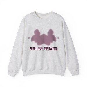 May include: A light grey sweatshirt with a crew neck. The front displays a purple ink blot design and the text "ERROR 404: MOTIVATION" in a dark purple font. Below the text is the phrase "I'M WATCHING YOU WATCH ME".