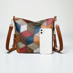 Peut inclure: Sac bandoulière patchwork multicolore avec un motif géométrique. Le sac présente des carrés de cuir dans les tons de bleu, rouge, orange et marron. Il possède des lanières et des pompons en cuir marron, et une chaîne dorée. Un smartphone blanc est placé devant le sac.