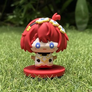 Könnte beinhalten: Kleine Figur mit rotem Haar, einem Blumenstirnband und einem weiß-goldenen Outfit. Die Figur hat große blaue Augen und steht auf einem roten kreisförmigen Sockel. Der Hintergrund ist grünes Gras.