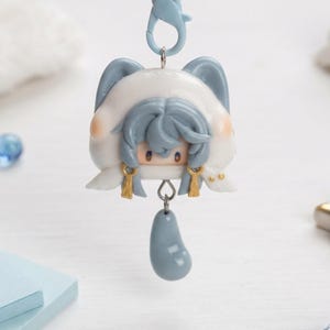 May include: A keychain featuring a sculpted character head with blue hair and a white hood with blue ears. The figure has small gold-colored accents and a dangling blue teardrop-shaped charm attached by a silver metal ring and a blue plastic clasp.
