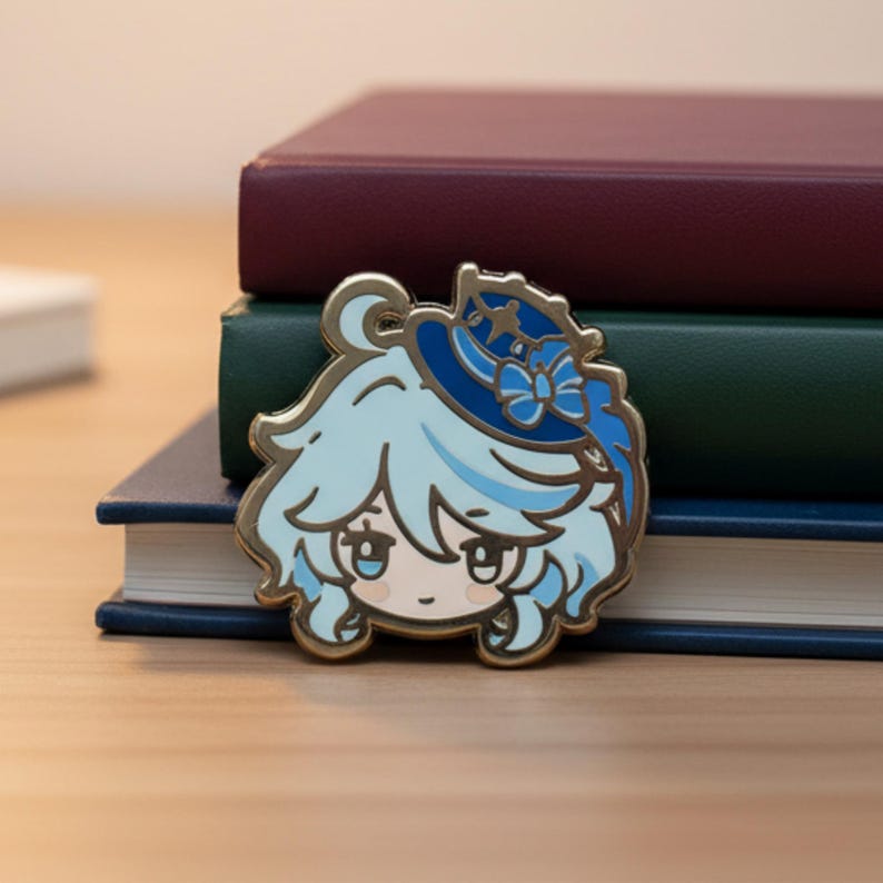Peut inclure: &Eacute;pingle en &eacute;mail dor&eacute;e repr&eacute;sentant un personnage de dessin anim&eacute; aux cheveux bleu clair et un chapeau bleu avec un n&oelig;ud. L'&eacute;pingle est pos&eacute;e sur un fond de livres empil&eacute;s avec des couvertures bordeaux, vertes et bleues.