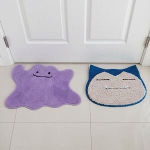 May include: Two character-shaped floor mats. One is purple and blob-like with a simple smiling face. The other is blue and beige, resembling a sleeping cartoon character. Both mats are on a tiled floor in front of a white door.