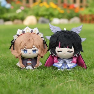 May include: Two plush dolls with detailed outfits and hairstyles are displayed on green grass. One doll has brown hair and a decorative headpiece, while the other has black hair with pink accents and small wings.