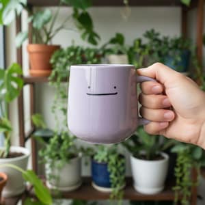 Könnte beinhalten: Eine lavendelfarbene Keramiktasse mit einem schwarzen Smiley-Gesicht-Design. Die Tasse hat eine abgerundete Form und einen passenden lavendelfarbenen Griff. Der Hintergrund zeigt eine Vielzahl von Topfpflanzen auf Holzregalen.