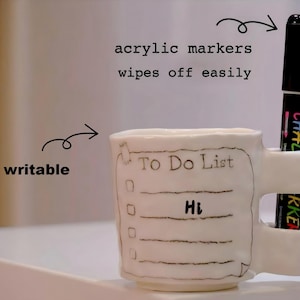 May include: A white ceramic mug with a "To Do List" design and a black marker. The mug has a handle and a slot to hold the marker. The text "Hi" is written on the mug. The image also includes the text "acrylic markers wipes off easily" and "writable".