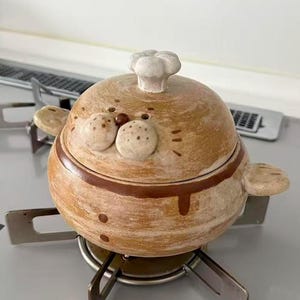 May include: A round, tan ceramic pot with a lid featuring a sculpted seal face and a small chef hat knob. The pot has two side handles, a brown horizontal band around the middle, and speckled details on the seal's snout and whiskers.