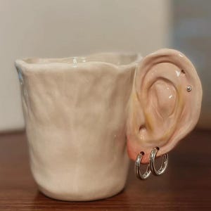 Puede incluir: Una taza de cerámica blanca con superficie texturizada. El asa tiene forma de oreja humana, con aros plateados y un pendiente. La oreja está pintada en un tono de piel.