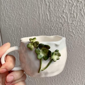 May include: White ceramic mug with a textured surface and a curved handle. The mug is adorned with three-dimensional green shamrock accents. The mug is held by a hand.