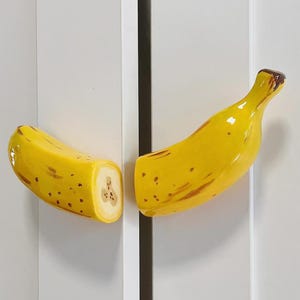 Peut inclure: Deux poignées de placard en céramique en forme de banane. Les poignées sont jaunes avec des taches marron et une tige marron. L'une des poignées est coupée pour montrer l'intérieur de la banane. Les poignées sont montées sur une porte d'armoire blanche.