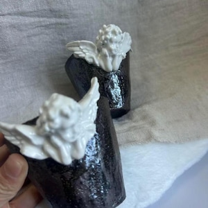 Puede incluir: Dos tazas de cer&aacute;mica con un exterior oscuro y texturizado y una figura de querub&iacute;n blanca en la parte superior. Los querubines tienen alas detalladas y cabello rizado. Las tazas est&aacute;n sobre un fondo neutro.