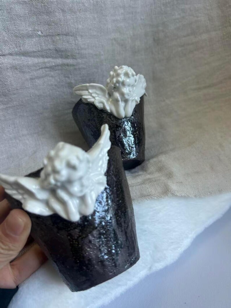 Puede incluir: Dos tazas de cer&aacute;mica con un exterior oscuro y texturizado y una figura de querub&iacute;n blanca en la parte superior. Los querubines tienen alas detalladas y cabello rizado. Las tazas est&aacute;n sobre un fondo neutro.