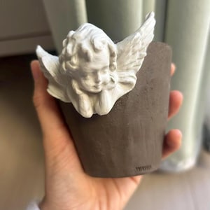 Puede incluir: Una peque&ntilde;a maceta de cer&aacute;mica marr&oacute;n con una figura de &aacute;ngel querub&iacute;n blanco adjunta. El &aacute;ngel tiene alas detalladas y una expresi&oacute;n serena. La maceta se sostiene en una mano, con un fondo borroso.