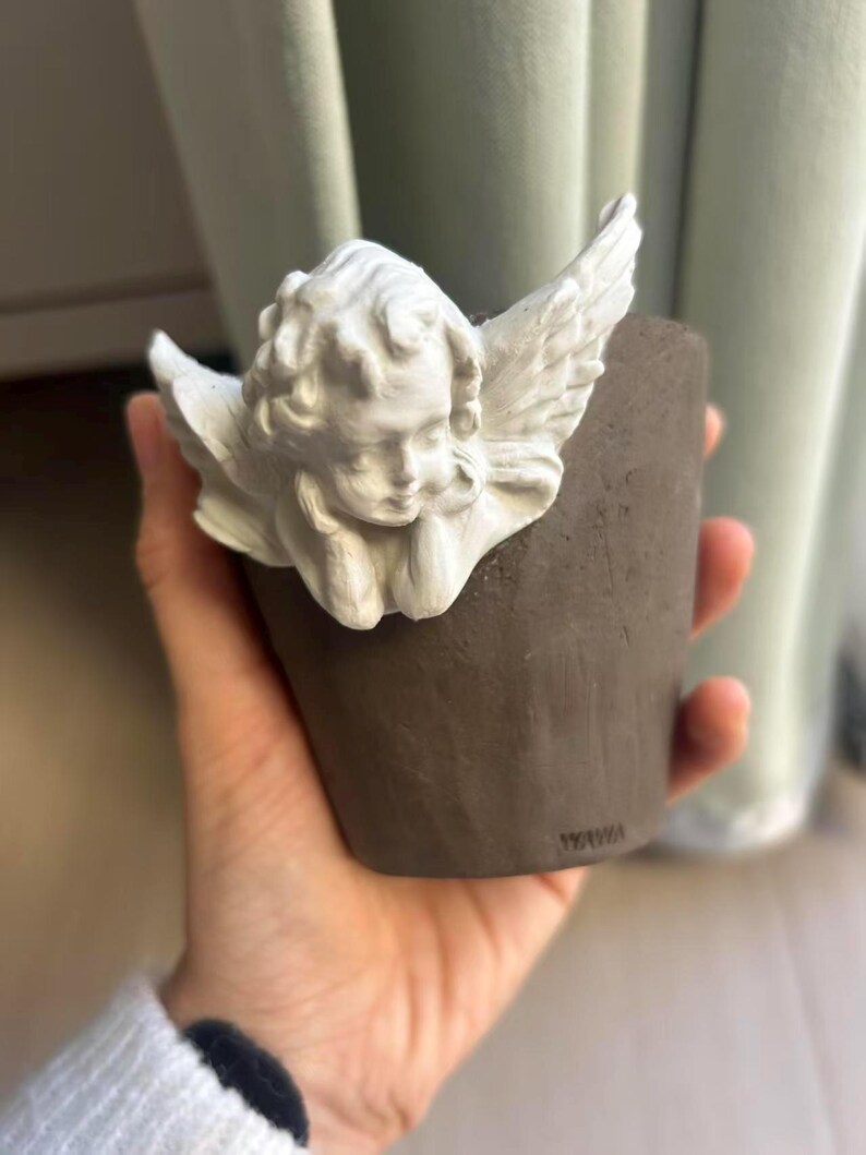 Puede incluir: Una peque&ntilde;a maceta de cer&aacute;mica marr&oacute;n con una figura de &aacute;ngel querub&iacute;n blanco adjunta. El &aacute;ngel tiene alas detalladas y una expresi&oacute;n serena. La maceta se sostiene en una mano, con un fondo borroso.