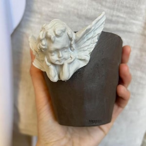 Puede incluir: Una peque&ntilde;a maceta de cer&aacute;mica marr&oacute;n oscuro con una figura de &aacute;ngel querub&iacute;n blanco adjunta. El &aacute;ngel tiene la cabeza apoyada en la mano y tiene alas detalladas. La maceta se sostiene en una mano.