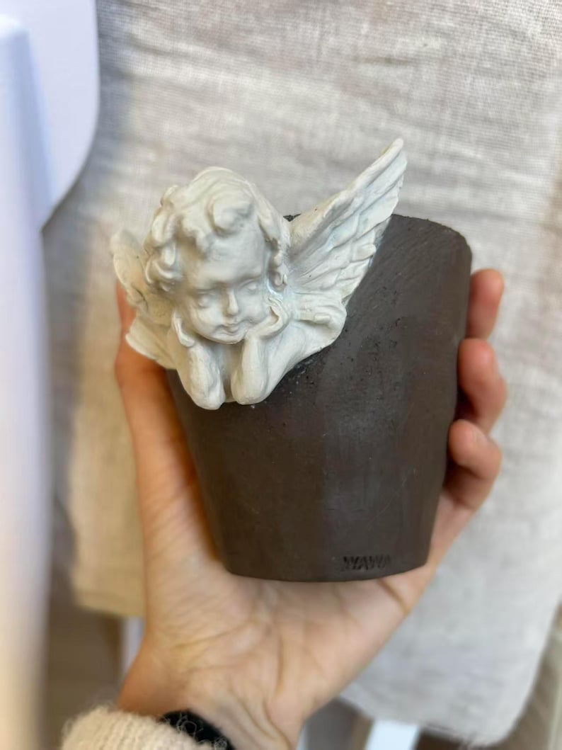 Puede incluir: Una peque&ntilde;a maceta de cer&aacute;mica marr&oacute;n oscuro con una figura de &aacute;ngel querub&iacute;n blanco adjunta. El &aacute;ngel tiene la cabeza apoyada en la mano y tiene alas detalladas. La maceta se sostiene en una mano.