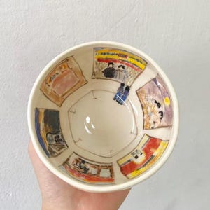 May include: A cream-colored ceramic bowl with hand-painted illustrations. The bowl's interior features a series of colorful, abstract scenes. The rim is a simple, unadorned design. The bowl is held by a person.
