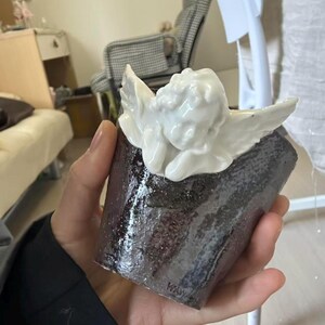 Puede incluir: Una taza de cer&aacute;mica hecha a mano con un exterior oscuro y texturizado y una figura de querub&iacute;n blanca adjunta en la parte superior. El querub&iacute;n tiene alas detalladas y est&aacute; acostado. La taza se sostiene en una mano.