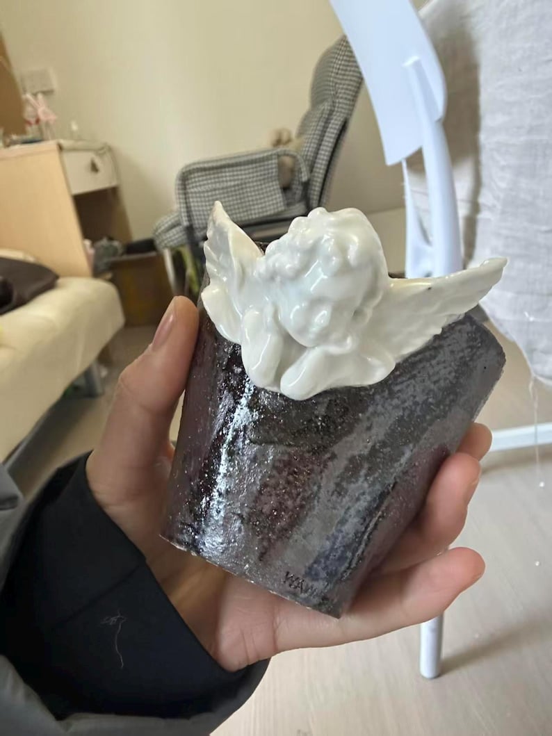 Puede incluir: Una taza de cer&aacute;mica hecha a mano con un exterior oscuro y texturizado y una figura de querub&iacute;n blanca adjunta en la parte superior. El querub&iacute;n tiene alas detalladas y est&aacute; acostado. La taza se sostiene en una mano.