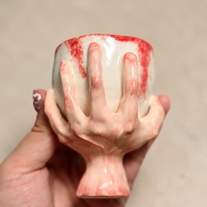 Puede incluir: Una taza de cerámica con una mano esculpida envuelta alrededor de la base y los lados exteriores. La taza tiene un acabado brillante en color blanco roto con detalles realistas en tono piel en la mano y salpicaduras de color rojo vivo.