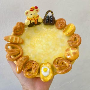 Puede incluir: Un plato decorativo de cerámica amarilla con un borde de productos horneados en miniatura, que incluyen pretzels, pan y panecillos. El plato presenta una figura de oso con una bufanda y un pequeño bolso. El plato está sostenido por una mano.