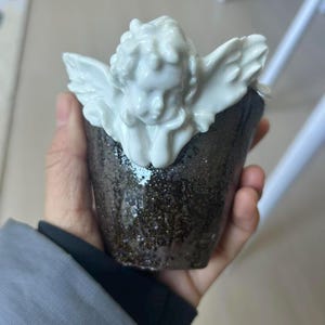 Puede incluir: Una taza de cer&aacute;mica con una figura de querub&iacute;n blanco en la parte superior. La taza tiene un exterior oscuro y moteado. El querub&iacute;n tiene alas y rasgos faciales detallados. La taza est&aacute; en la mano de una persona.
