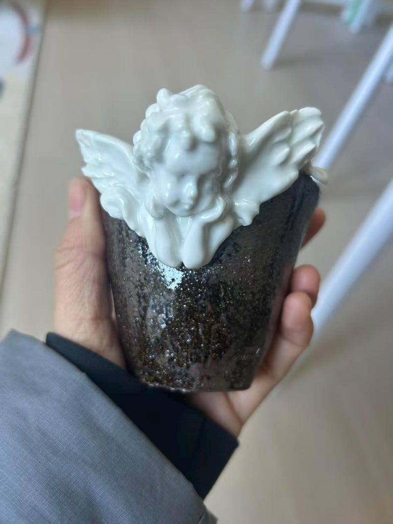 Puede incluir: Una taza de cer&aacute;mica con una figura de querub&iacute;n blanco en la parte superior. La taza tiene un exterior oscuro y moteado. El querub&iacute;n tiene alas y rasgos faciales detallados. La taza est&aacute; en la mano de una persona.