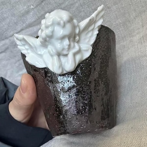 Puede incluir: Una peque&ntilde;a taza de cer&aacute;mica marr&oacute;n oscuro con una figura de querub&iacute;n blanco unida al borde. El querub&iacute;n tiene la cabeza apoyada en sus manos y tiene alas detalladas. La taza tiene una superficie texturizada y una forma ligeramente irregular.