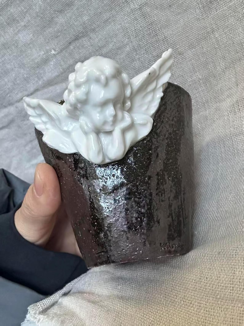 Puede incluir: Una peque&ntilde;a taza de cer&aacute;mica marr&oacute;n oscuro con una figura de querub&iacute;n blanco unida al borde. El querub&iacute;n tiene la cabeza apoyada en sus manos y tiene alas detalladas. La taza tiene una superficie texturizada y una forma ligeramente irregular.