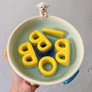 Op de afbeelding: Een keramische kom met een lichtblauwe binnenkant en een witte rand. De kom bevat gele lettervormige objecten en is bekroond met een klein wit hondenbeeldje. De kom heeft kleurrijke handvatten.