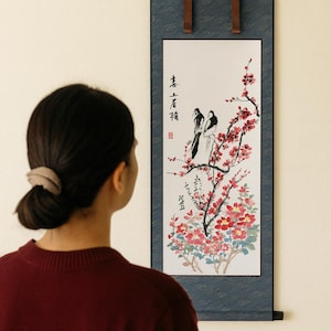 May include: A vertical scroll painting featuring a watercolor illustration of birds perched on a flowering branch with red blossoms. The artwork is framed by a dark blue border and brown leather straps.