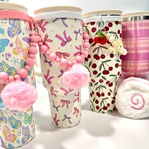 May include: Several insulated tumblers with various designs are displayed. One features butterflies, another has pink bows, and a third showcases cherries. Each tumbler has a white rim and is adorned with accessories like a pink pom-pom.