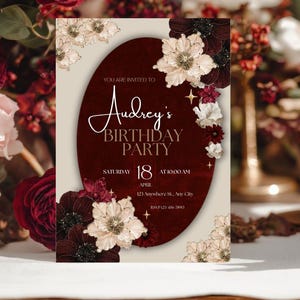 May include: A birthday party invitation with a burgundy oval in the center, adorned with floral accents. The invitation reads "Audrey's Birthday Party" with the date, time, and location details. The design features a cream background and elegant script.