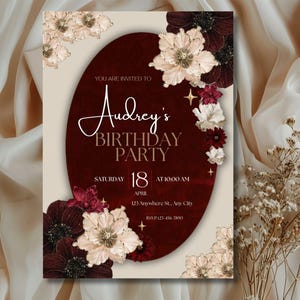 May include: A birthday party invitation with a burgundy oval in the center, adorned with floral accents. The invitation reads "Audrey's Birthday Party" with the date, time, and location details. The design features cream and burgundy colors.