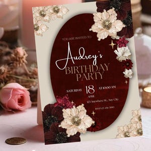 May include: A birthday party invitation with a burgundy oval design, featuring the text "Audrey's BIRTHDAY PARTY". The invitation includes the date, time, and location. Floral accents in cream, burgundy, and white decorate the corners.