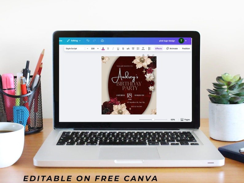 May include: A laptop displays a birthday party invitation design. The invitation features a burgundy oval with floral accents and the text "Audrey's Birthday Party." The laptop is on a wooden desk with office supplies.