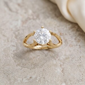 Peut inclure: Une bague en or avec une pierre ronde transparente taille brillant, fixée par un sertissage à six griffes. L'anneau présente un design divisé qui s'incurve vers le haut pour rejoindre la pierre centrale, créant un aspect ouvert et symétrique.