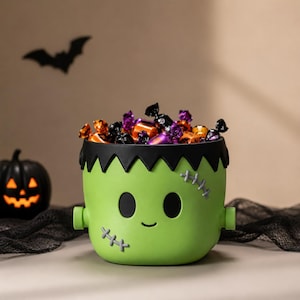 Puede incluir: Un cubo de color verde claro con forma de cabeza de monstruo, con pelo negro, dos ojos ovalados negros, una pequeña sonrisa y puntadas grises en las mejillas. El cubo está lleno de caramelos variados en envoltorios naranjas, morados y negros.