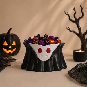 May include: A ceramic bowl shaped like a vampire with a white face, red eyes, and fangs, wearing a black cape. The bowl is filled with assorted candies wrapped in orange, purple, and black foil, creating a festive display for the Halloween season.