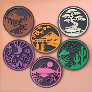 May include: Set of six round coasters with nature-themed designs. Each coaster features a different landscape, including mountains, a bonsai tree, and a desert scene with a cactus. The coasters are in various colors, including brown, orange, green, purple, and pink.
