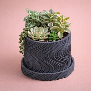 Può includere: Un vaso da fiori grigio scuro, strutturato, con un design ondulato, riempito con varie piante succulente e piante pendenti. Il vaso include un sottovaso abbinato. Le piante presentano sfumature di verde e bianco.