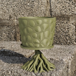 Puede incluir: Una maceta verde con un diseño texturizado similar a guijarros. La maceta se apoya en un platillo a juego, sostenido por una base con forma de raíces entrelazadas. Un artículo decorativo de inspiración natural para plantas.