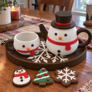 Puede incluir: Una tetera blanca con forma de muñeco de nieve, con sombrero de copa marrón, bufanda roja y asa marrón, junto a una taza a juego sobre una bandeja redonda marrón con un diseño de copo de nieve blanco. Tres adornos planos con forma de muñeco de nieve, árbol y copo de nieve se sitúan delante.