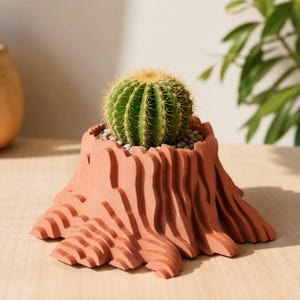 May include: A green cactus with yellow spines sits in a terracotta-colored planter shaped like a tree stump. The planter has a textured, layered design. Small pebbles surround the base of the cactus.