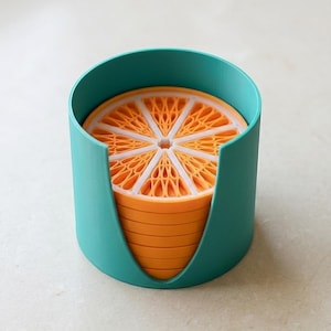 Puede incluir: Un conjunto de posavasos en forma de rodaja de naranja con detalles blancos y naranjas, guardados en un soporte turquesa. Los posavasos están apilados ordenadamente dentro del soporte, creando un diseño vibrante y juguetón.