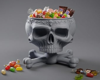 May include: A matte gray 3D-printed skull-shaped bowl featuring an intricate scrollwork border with a small skull detail at the center. The bowl is supported by a base formed from two crossed bones and is filled with assorted colorful candies.
