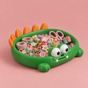 May include: A green monster-shaped storage tray with orange spikes along the back, two large white eyes, and two small white fangs. The tray is filled with various craft supplies, including colorful beads, small pink scissors, patterned tape rolls, and buttons.